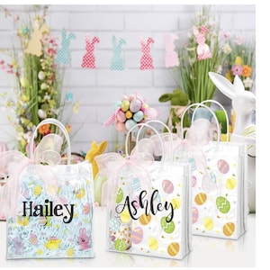 May include: Clear Easter gift bags with white handles and pink bows. The bags are decorated with colorful Easter eggs and the names "Hailey" and "Ashley" in black script. The bags are filled with blue shredded paper and Easter-themed toys.