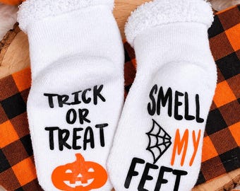 May include: A pair of white fuzzy ankle socks with black and orange text and graphics. The left sock features the text "TRICK OR TREAT" with an orange pumpkin. The right sock features the text "SMELL MY FEET" with a black spiderweb graphic.
