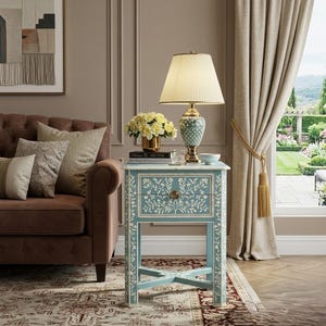May include: A light blue bedside table with white floral inlay patterns, a single drawer with a round knob, and an X-shaped base. On top sits a table lamp with a pleated cream shade and a patterned base, alongside a vase of yellow flowers.
