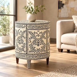 May include: A cylindrical side table featuring a cream-colored surface with intricate black floral and vine patterns. The table has a single door with a small round knob, supported by three tapered legs, and is topped with a round vase.
