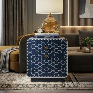 May include: A navy blue bedside table with a white hexagonal honeycomb pattern, featuring two circular gold knobs. On top sits a glass table lamp with a white shade, a small analog clock, and two books titled Roman Pol and Mixestane.