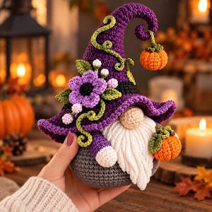 May include: A crochet gnome featuring a tall purple pointed hat adorned with green vines, small white flowers, and a large purple flower. The gnome has a white beard, a beige nose, and a gray body, with small orange pumpkins attached to the hat and side.