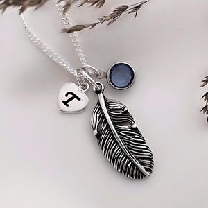 May include: A silver necklace with a feather charm, a heart charm with the letter "T" engraved on it, and a small blue gemstone charm.