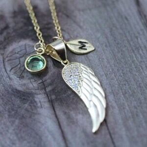 May include: Gold necklace with a green gemstone, a small leaf charm with the letter "M" and a silver wing charm with clear rhinestones.