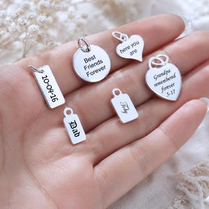 Könnte beinhalten: Eine Sammlung von sechs silberfarbenen Metallanhängern in verschiedenen Formen wie Herzen, Kreisen und Rechtecken. Jeder Anhänger ist mit Texten graviert, darunter Best Friends Forever, here you are, 10-04-16, Dad, Toby und Grandpa remembered forever 5-17.
