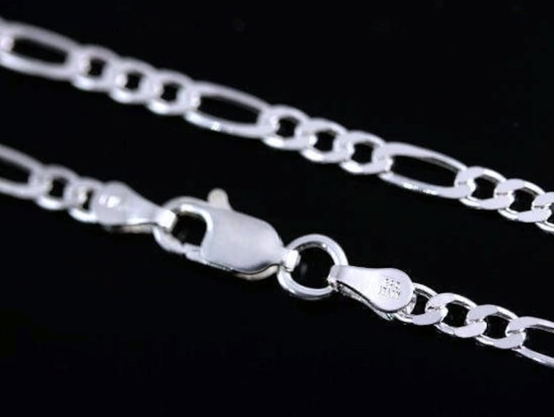 May include: A silver Figaro chain necklace featuring a polished finish and a lobster claw clasp. The metal tag near the clasp is stamped with the text 925 and ITALY, indicating the material composition and origin of the jewelry piece.
