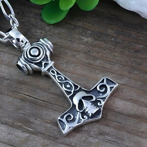 May include: A silver-toned metal pendant shaped like a hammer, featuring an engraved skull design on the head and intricate swirling patterns along the handle. The pendant is attached to a silver-toned metal chain with oval-shaped links.