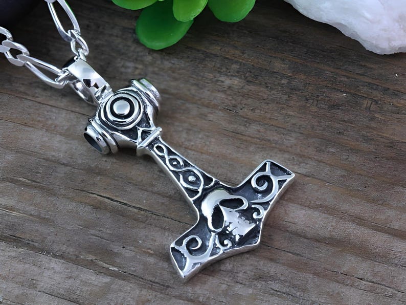 May include: A silver-toned metal pendant shaped like a hammer, featuring an engraved skull design on the head and intricate swirling patterns along the handle. The pendant is attached to a silver-toned metal chain with oval-shaped links.