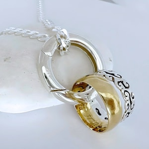 Puede incluir: Un collar de plata con un gran anillo de plata pulida y una banda dorada. La banda dorada tiene un diseño calado y una superficie lisa. La cadena del collar es una fina cadena de plata. La banda dorada está entrelazada con el anillo de plata.