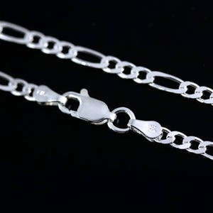 May include: A silver-toned Figaro chain necklace featuring a series of flattened circular links interspersed with elongated oval links. The chain is secured with a polished lobster claw clasp mechanism, showing clear metal link construction.