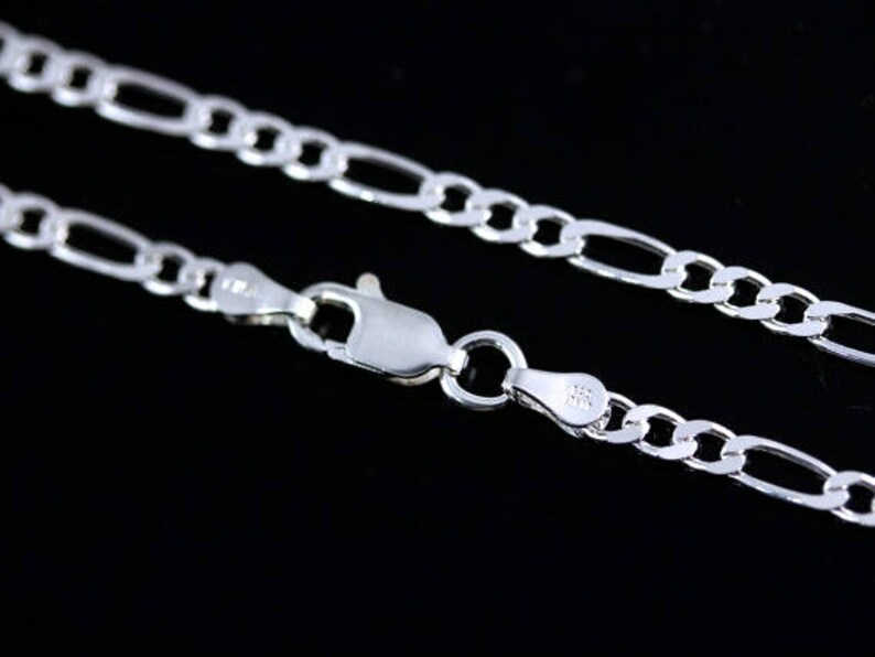 May include: A silver-toned Figaro chain necklace featuring a series of flattened circular links interspersed with elongated oval links. The chain is secured with a polished lobster claw clasp mechanism, showing clear metal link construction.