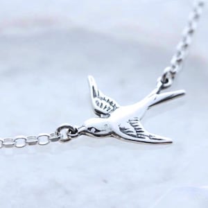 May include: A silver-colored necklace featuring a detailed bird pendant in flight. The bird has outstretched wings and is attached to a delicate chain. The necklace is displayed on a light-colored surface, highlighting the jewelry's design.