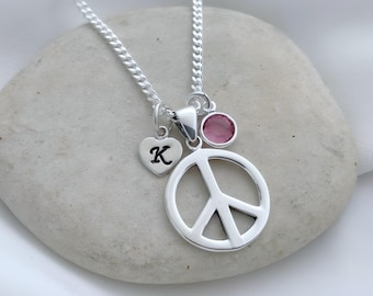 May include: A silver-toned necklace featuring a circular peace sign pendant, a small heart-shaped charm engraved with the letter K, and a round pink crystal charm, all suspended from a delicate silver-toned chain link necklace.