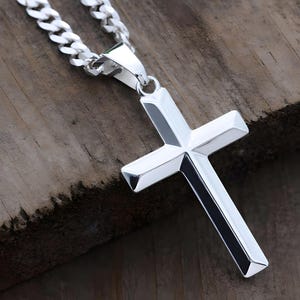 Op de afbeelding: Zilveren kruis hanger met bijpassende ketting. Het kruis heeft een gepolijst, reflecterend oppervlak en een eenvoudig ontwerp. De ketting is een curb link stijl. De hanger wordt gepresenteerd op een houten oppervlak.