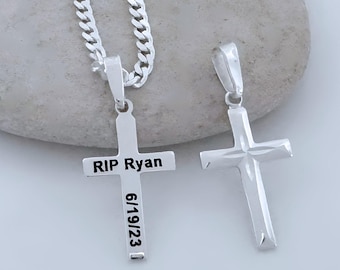 May include: Two silver-toned cross pendants, one attached to a curb chain. The left cross features the engraved text "RIP Ryan" horizontally and "6/19/23" vertically. The right cross has a plain, polished surface with a faceted center design.