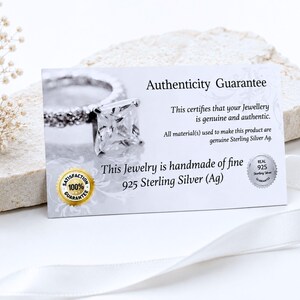 May include: A rectangular white authenticity guarantee card featuring a photograph of a silver ring with a square stone. The text reads Authenticity Guarantee, This certifies that your Jewellery is genuine and authentic, and 925 Sterling Silver.