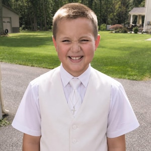 May include: A short-sleeved white button-down shirt paired with a cream-colored vest and a matching cream necktie. A thin silver chain with a small silver cross pendant is worn around the neck, resting over the center of the necktie.