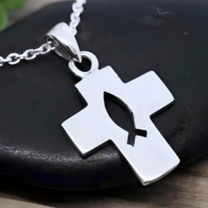 Peut inclure: Un pendentif croix en métal argenté suspendu à une chaîne assortie. La croix présente une découpe centrale en forme de symbole de poisson, créant un design en espace négatif au sein de la surface métallique solide, lisse et polie.