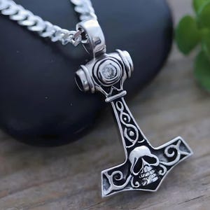 May include: A silver-toned metal pendant shaped like a hammer, featuring an engraved skull design at the base and intricate scrollwork patterns along the handle. The pendant is attached to a silver-toned curb chain necklace with a polished finish.