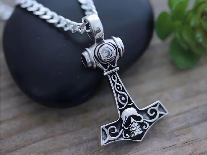 May include: A silver-toned metal pendant shaped like a hammer, featuring an engraved skull design at the base and intricate scrollwork patterns along the handle. The pendant is attached to a silver-toned curb chain necklace with a polished finish.