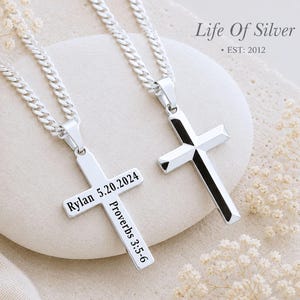 May include: Two silver-toned cross pendants on matching curb chains. One cross is plain and polished, while the other is engraved with the text Rylan 5.20.2024 and Proverbs 3:5-6. The pendants are displayed together on a smooth, rounded surface.