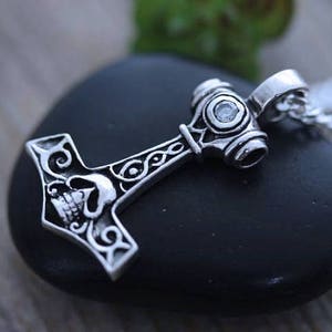May include: A silver-toned metal pendant shaped like a hammer, featuring an engraved skull design on the head and intricate scrollwork patterns along the handle. A small, clear, circular gemstone is set into the top of the hammer head.