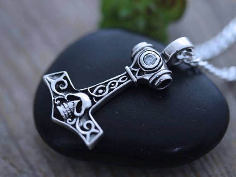 May include: A silver-toned metal pendant shaped like a hammer, featuring an engraved skull design on the head and intricate scrollwork patterns along the handle. A small, clear, circular gemstone is set into the top of the hammer head.