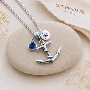 Puede incluir: Un collar con colgante de ancla en tono plateado que presenta un pequeño dije circular grabado con la letra M y un dije de piedra azul redonda. El collar incluye una delicada cadena plateada y se muestra con el texto LIFE OF SILVER EST. 2012.