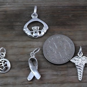 May include: Four silver-toned metal charms including a Claddagh, a circular Om symbol, a awareness ribbon, and a caduceus symbol. A United States dime is included for scale, showing the charms are approximately 0.7 inches in diameter or smaller.
