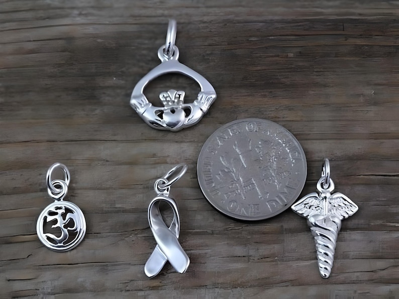 May include: Four silver-toned metal charms including a Claddagh, a circular Om symbol, a awareness ribbon, and a caduceus symbol. A United States dime is included for scale, showing the charms are approximately 0.7 inches in diameter or smaller.