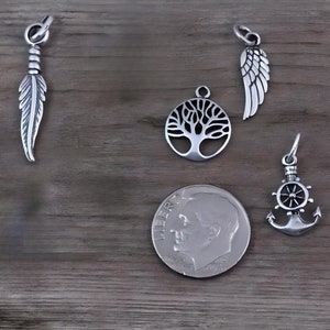 May include: Four silver-toned metal charms including a feather, a tree of life, an angel wing, and a ship anchor with a steering wheel. A United States dime is placed next to the charms to provide a clear sense of scale for the jewelry pieces.