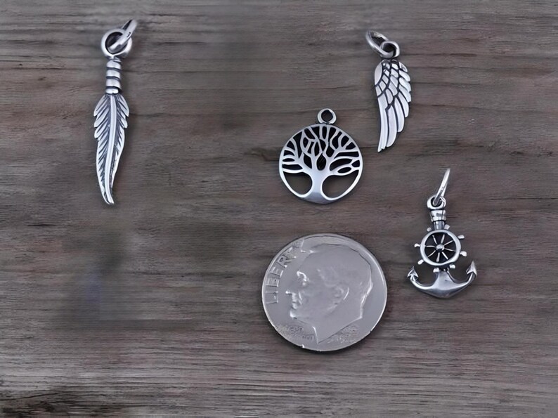 May include: Four silver-toned metal charms including a feather, a tree of life, an angel wing, and a ship anchor with a steering wheel. A United States dime is placed next to the charms to provide a clear sense of scale for the jewelry pieces.