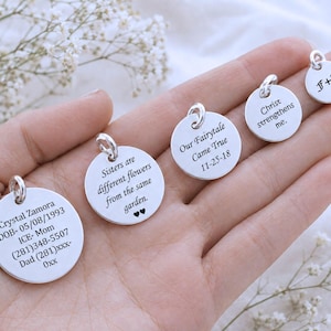 May include: Four circular silver charms with engraved text. The largest reads Crystal Zamora DOB- 05/08/1993 ICE- Mom (281)348-5507 Dad (281)xxx-0xx. Others read Sisters are different flowers from the same garden, Our Fairytale Came True 11-25-18, Christ strengthens me, and F+K.