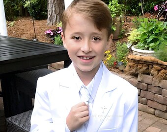 May include: A white blazer worn over a white collared shirt and a white tie, featuring a silver cross pendant on a chain. The blazer has a small silver cross pin attached to the left lapel area. The attire is formal, clean, and monochromatic.