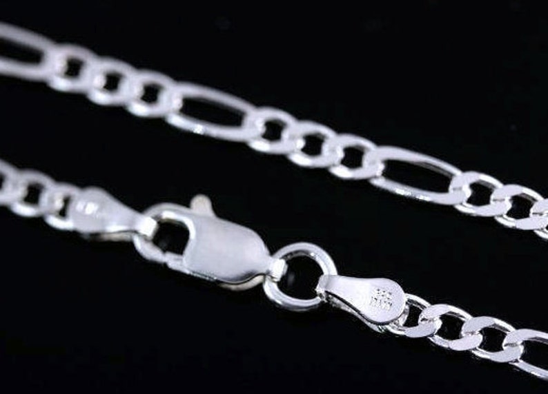 May include: A shiny silver figaro chain necklace featuring a polished finish and a secure lobster claw clasp. The metal links are arranged in a repeating pattern of three small circular links followed by one elongated oval link.