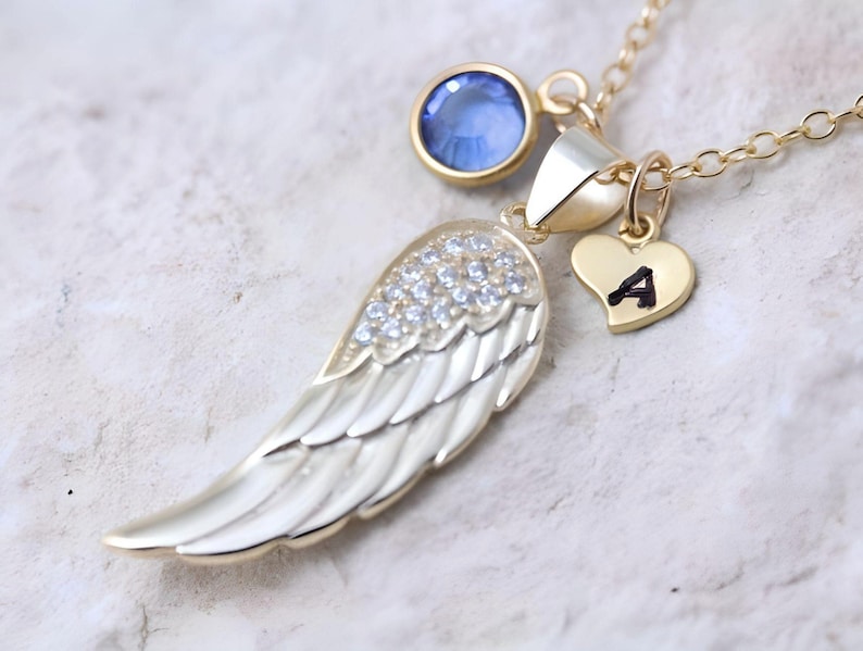 May include: Gold necklace with a small blue gemstone, a heart charm with the letter 'A' engraved on it, and a large angel wing pendant with clear rhinestones.