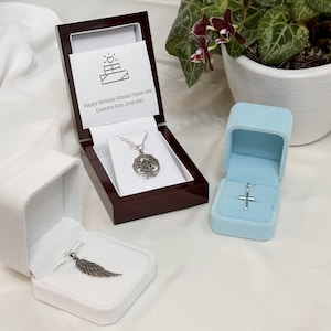 Puede incluir: Tres cajas de joyería de terciopelo blanco con collares de plata en su interior. Las cajas están sobre una superficie blanca. Los collares presentan un nudo celta, una cruz y un colgante de ala de ángel.