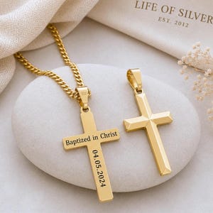 May include: Two gold-coloured cross pendants. One cross is on a gold chain and has the inscription "Baptized in Christ" and the date "04.05.2024". The other cross is a simple, solid design. The background includes a white stone and a fabric.