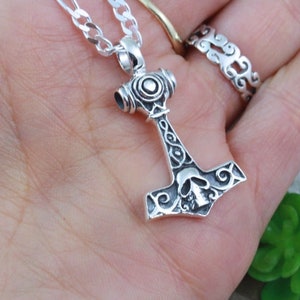 May include: A silver-colored metal pendant shaped like a hammer, featuring an engraved skull design on the head and intricate scrollwork patterns along the handle. The pendant is attached to a silver-colored chain link necklace.