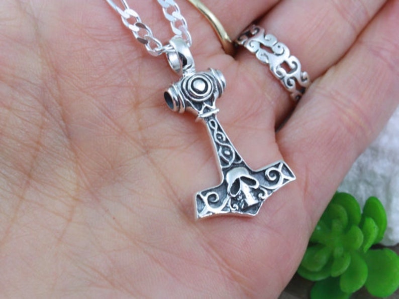 May include: A silver-colored metal pendant shaped like a hammer, featuring an engraved skull design on the head and intricate scrollwork patterns along the handle. The pendant is attached to a silver-colored chain link necklace.