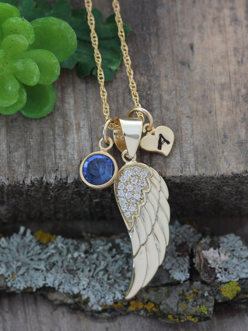 May include: Gold necklace with a blue gemstone, a small heart charm with the letter 'A' and a gold wing charm with clear crystals.