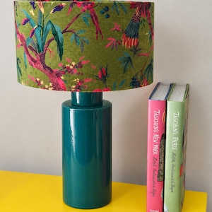 Puede incluir: Una lámpara de mesa con base cilíndrica de cerámica color verde azulado y una pantalla de tambor de tela verde con un patrón colorido de flores y pájaros. Dos libros titulados Taschen's New York y Taschen's Paris están junto a la base.