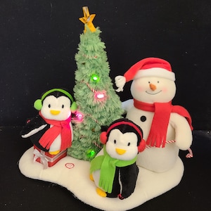 May include: A Christmas-themed plush display featuring a snowman wearing a red scarf and Santa hat, two penguins with earmuffs and scarves, and a green Christmas tree with lights. The display sits on a white base.