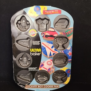 May include: A Wilton Ultra Bake 12-cavity boy cookie pan. The non-stick pan has molds for cookies shaped like a rocket, a sports jersey, a race car, a trophy, and more. The packaging includes the text "Improved Non-Stick".