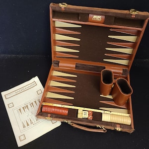 Puede incluir: Juego de backgammon abierto en un estuche de cuero marrón. El interior es marrón oscuro con triángulos beige y crema. El juego incluye dados, cubiletes y fichas. Se ve un papel con la palabra "BACKGAMMON".