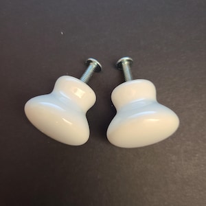 May include: Two white ceramic cabinet knobs with silver screws. The knobs have a smooth, glossy finish and a rounded, teardrop shape. They are set against a dark background.