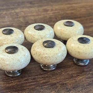 May include: A set of five round cabinet knobs featuring a cream-colored ceramic surface with small brown speckles. Each knob has a dark, weathered metal center inlay and a matching metallic base, providing a consistent look for furniture hardware.