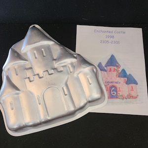 May include: A silver-colored, castle-shaped cake pan with a detailed design of turrets, windows, and a gate. A printed image of a pink castle with blue turrets and the text "Enchanted Castle 1998" is next to the pan.