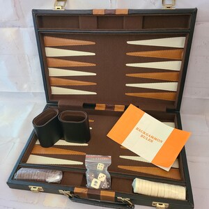May include: A black backgammon board game set in a briefcase-style case with a brown felt interior featuring tan and cream-colored triangular points. Includes two black dice cups, a stack of brown and cream checkers, dice, and a Backgammon Rules booklet.