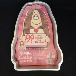 May include: A vintage Wilton Cathy Cake Pan. The silver-coloured pan is shaped like a woman with long hair. The insert features a cartoon image holding a sign that reads "Happy Birthday to a Real Hunk!"